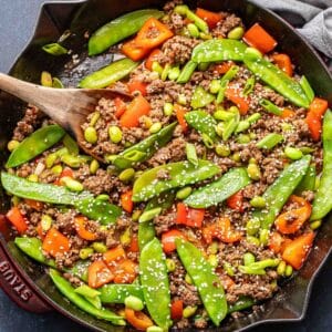 Ground beef stir fry with snow peas, red bell pepper, and edamame in a skillet with a wooden serving spoon.