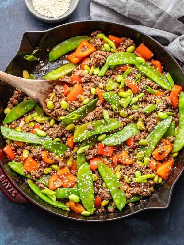 Ground beef stir fry with snow peas, red bell pepper, and edamame in a skillet with a wooden serving spoon.