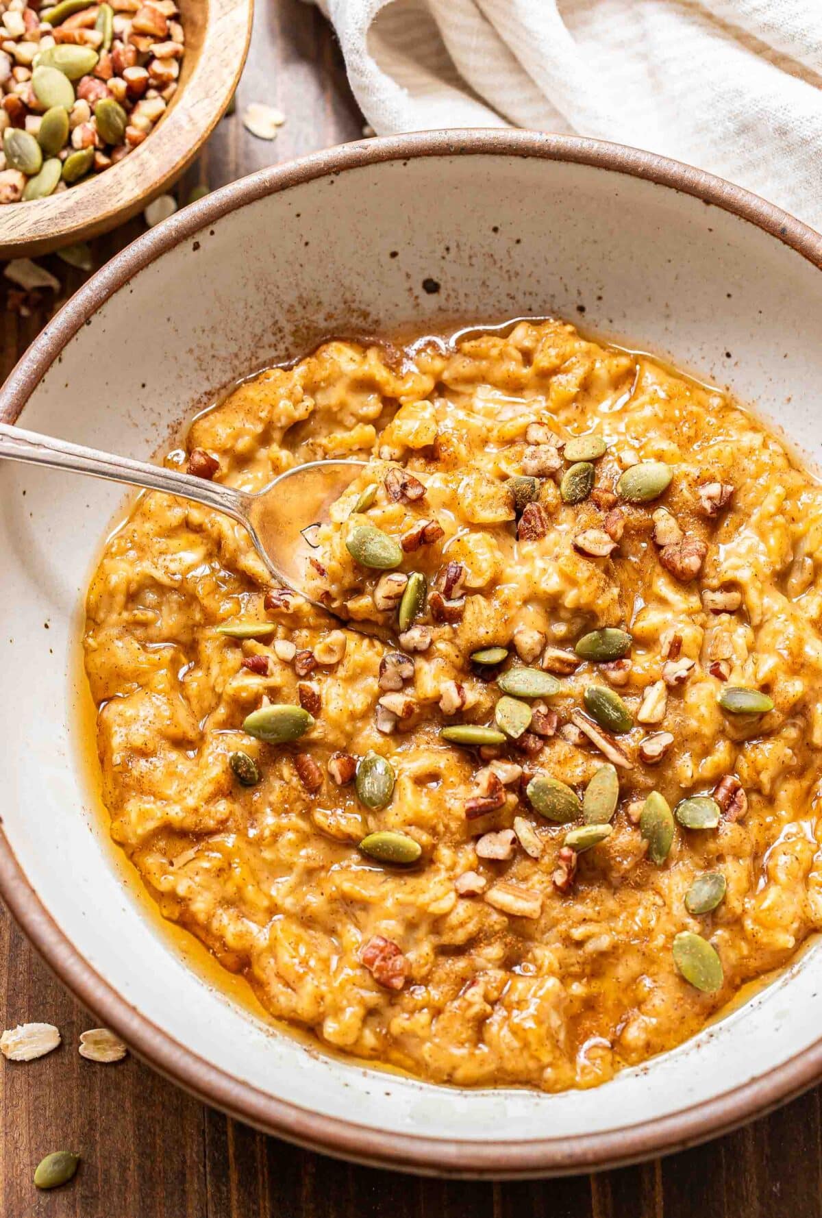 Pumpkin oatmeal in a white bowl with a spoon. The oatmeal is topped with pumpkin seeds, pecans, and maple syrup.