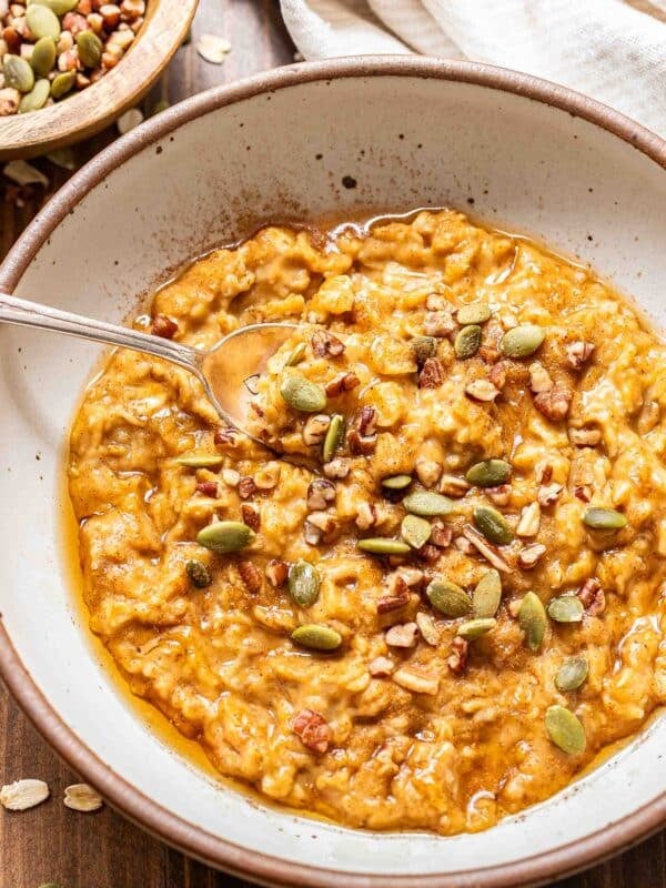 Pumpkin oatmeal in a white bowl with a spoon. The oatmeal is topped with pumpkin seeds, pecans, and maple syrup.