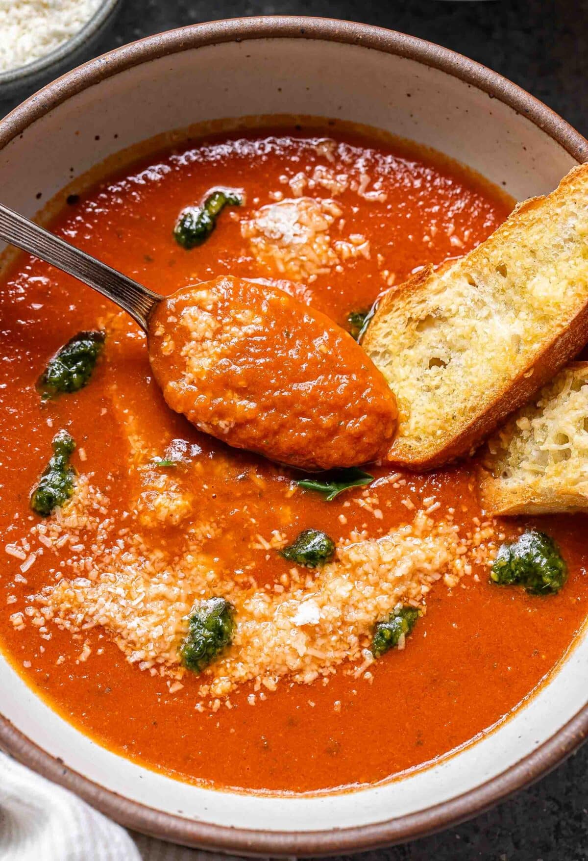 Tomato basil soup with pesto in a white bowl with a spoon and two slices of baguette.