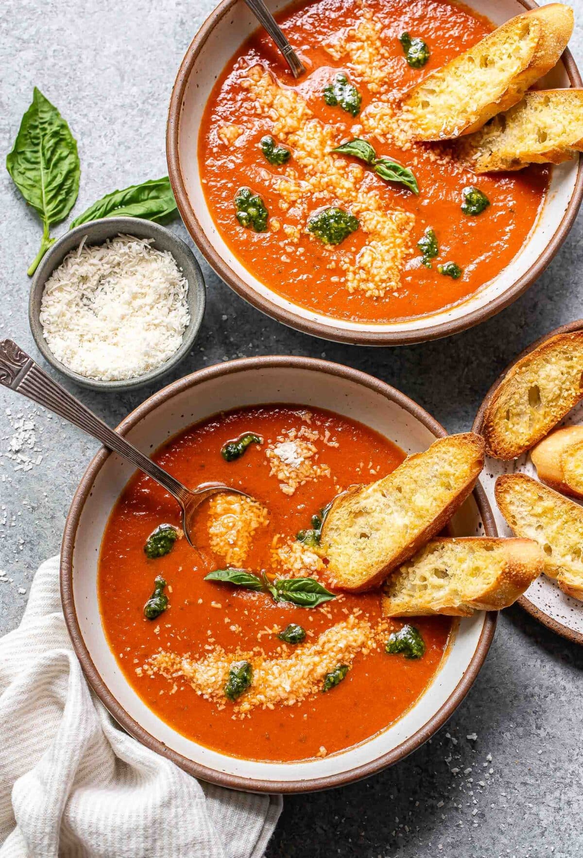 Two bowls of tomato basil soup topped with parmesan cheese. Two slices of toasted baguette are in each bowl.