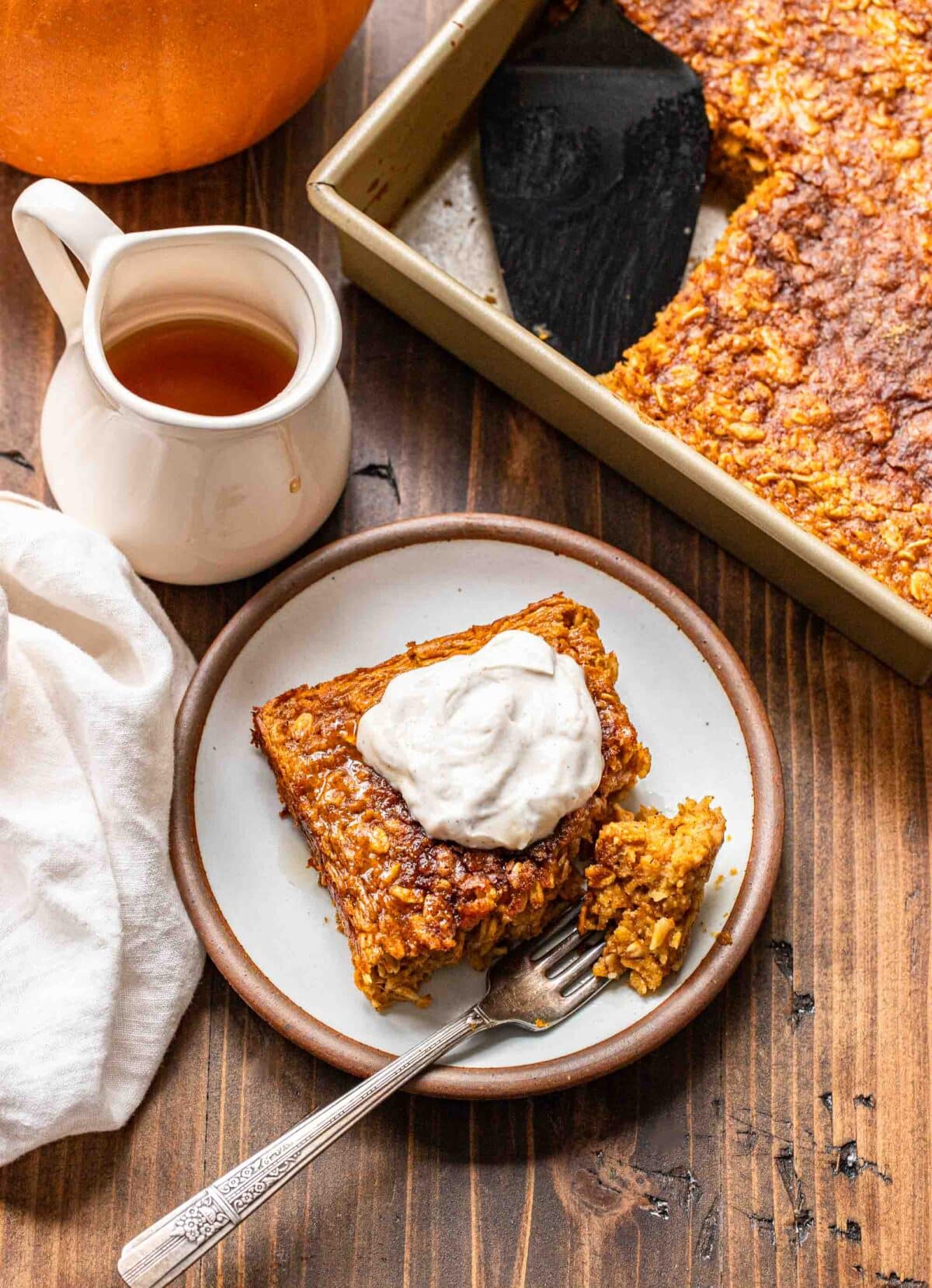 A piece of pumpkin baked oatmeal on a white plate with a fork. Maple syrup and the pan of baked oatmeal are behind the plate.