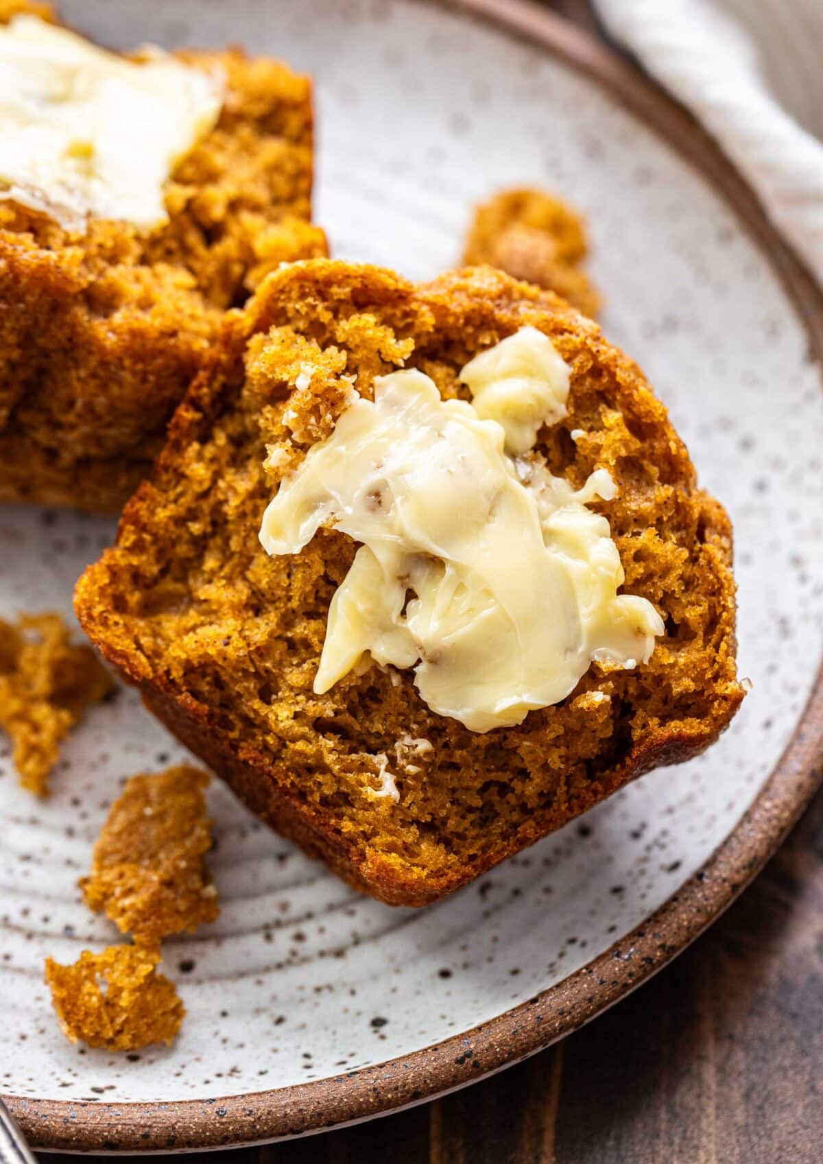 Half of a pumpkin muffin with butter on it on a white plate.