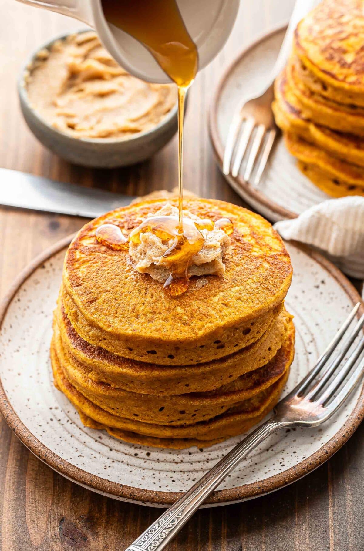 A stack of pumpkin pancakes on a white plate with maple syrup being poured onto them.