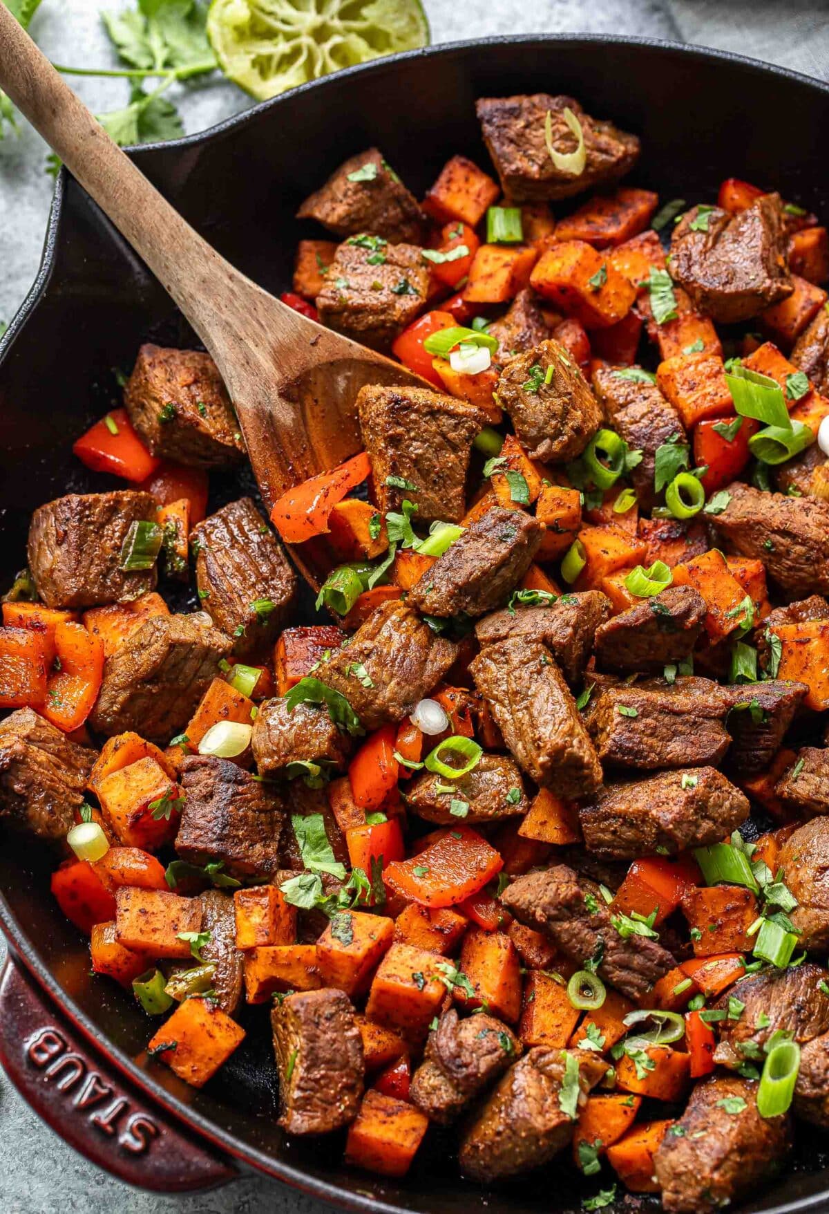 Chunks of steak, sweet potatoes, and red bell pepper in a skillet with a wooden spoon.