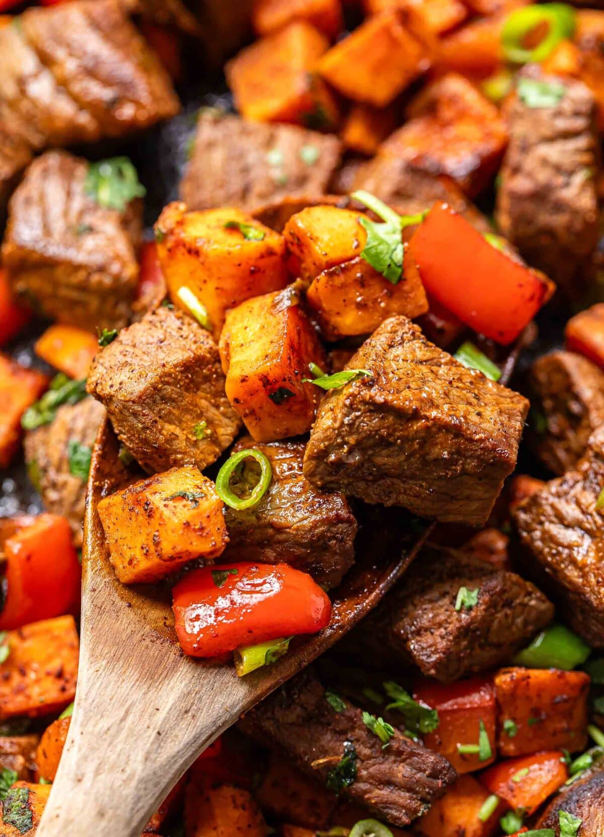 Chunks of steak, sweet potato and red bell pepper on a wooden spoon.