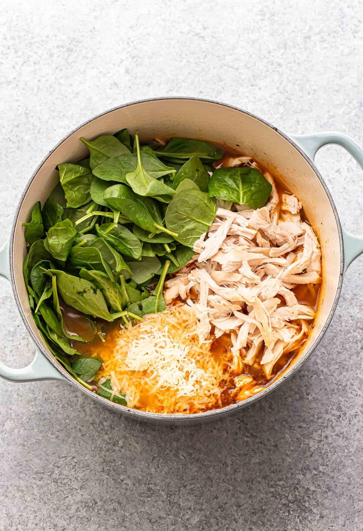 Soup broth in a pot with spinach, shredded cooked chicken, and shredded parmesan cheese.