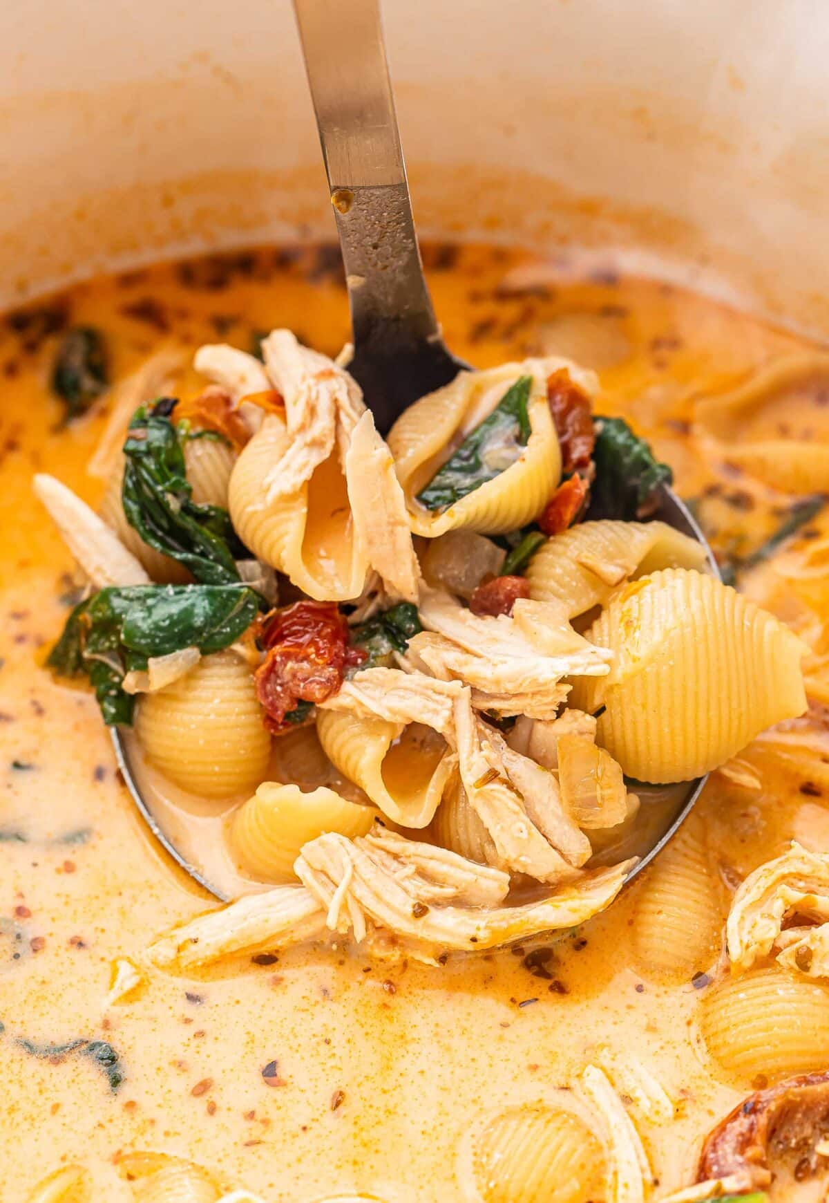 Ladle holding up a scoop of creamy soup with shredded chicken, spinach, and sun dried tomatoes.