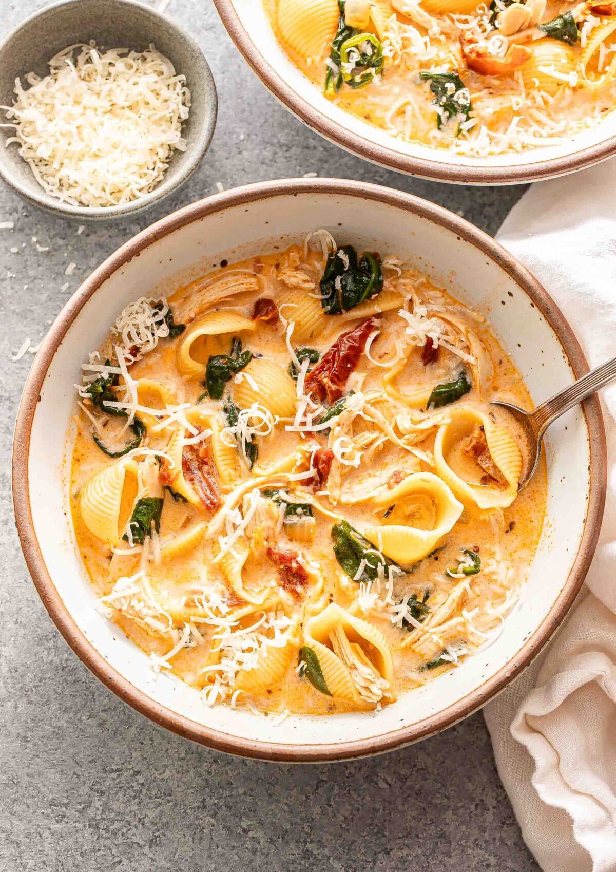 A white bowl with marry me chicken soup and a spoon in it. A bowl of parmesan cheese is behind the bowl.