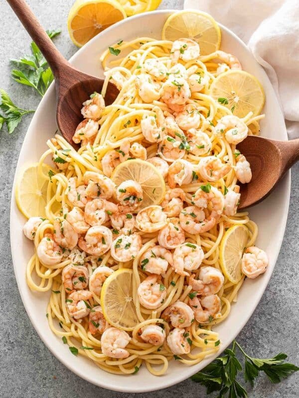 An oval white serving platter with pasta and shrimp scampi on it and two serving spoons.