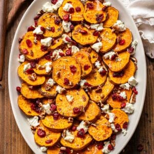 Roasted sweet potatoes on an oval platter with goat cheese crumbles, dried cranberries, and pecans.