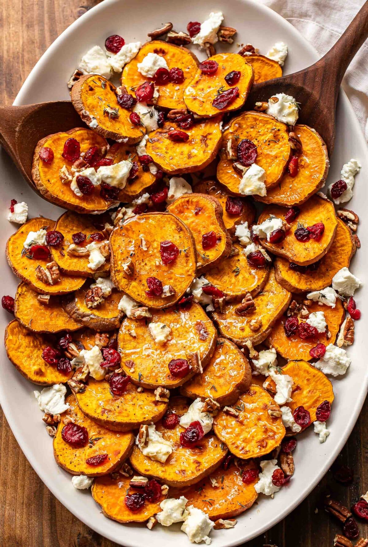 Roasted sweet potatoes on an oval platter with goat cheese crumbles, dried cranberries, and pecans and two wooden serving spoons.
