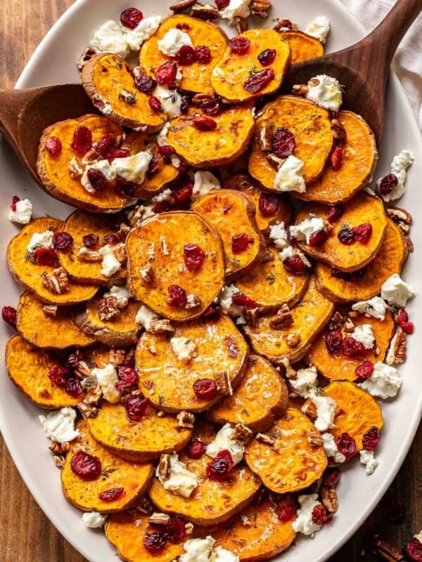 Roasted sweet potatoes on an oval platter with goat cheese crumbles, dried cranberries, and pecans and two wooden serving spoons.