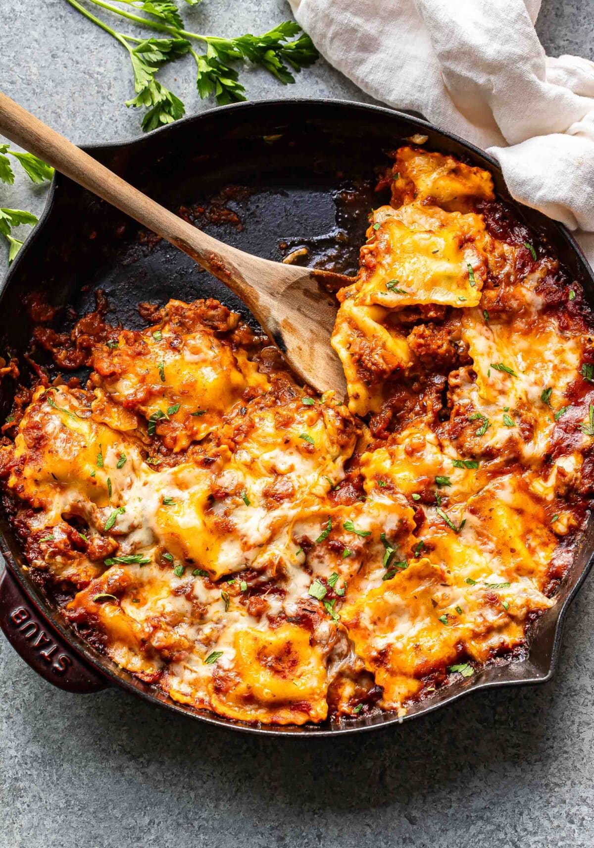Cheesy baked ravioli in marinara meat sauce in a skillet with a wooden serving spoon.