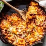 Cheesy baked ravioli in marinara meat sauce in a skillet with a wooden serving spoon.