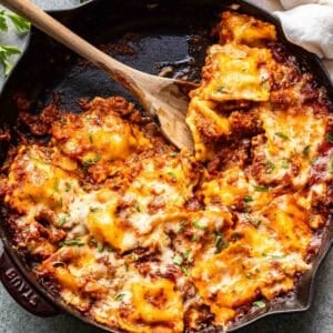 Cheesy baked ravioli in marinara meat sauce in a skillet with a wooden serving spoon.