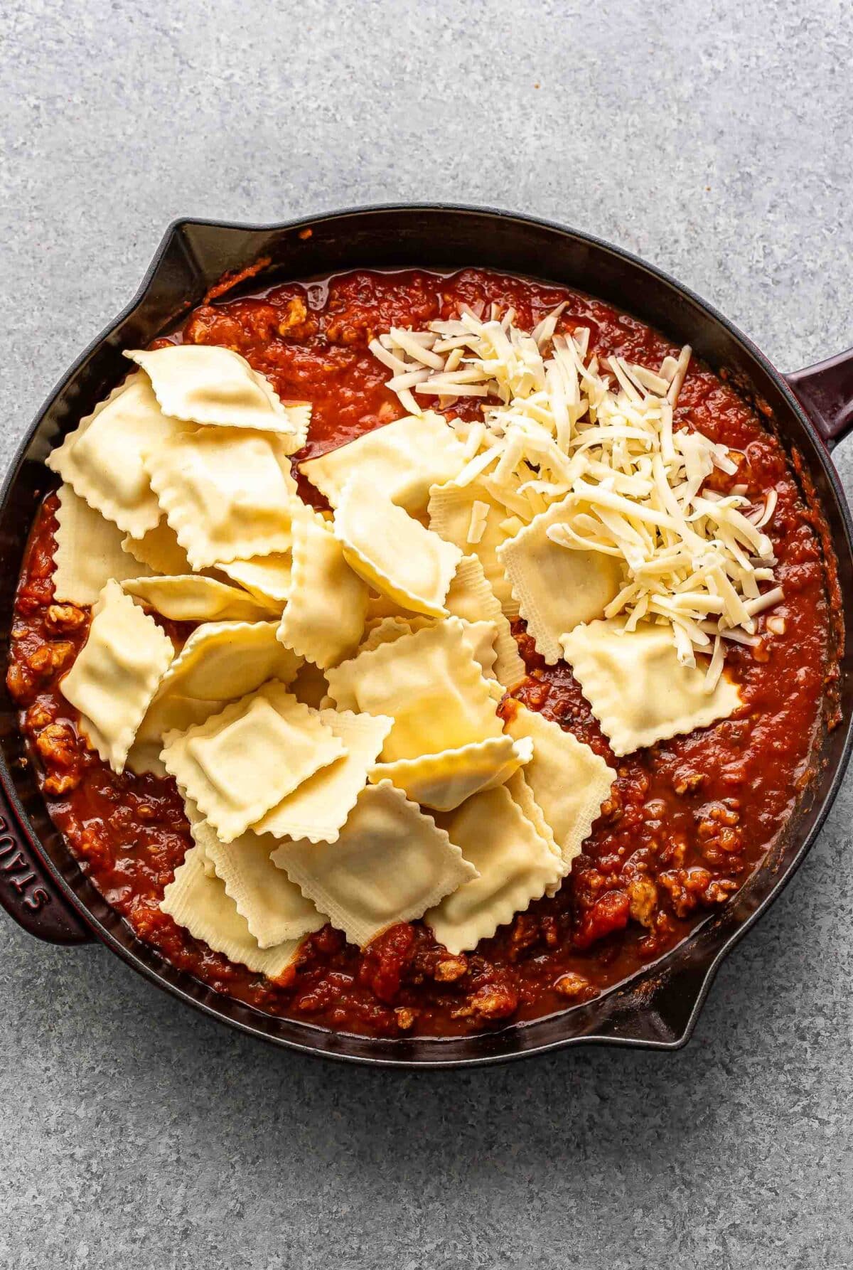 Marinara sauce with Italian sausage topped with ravioli and shredded cheese in a skillet.