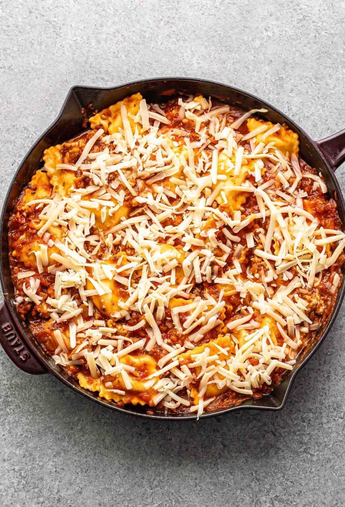 Ravioli covered in marinara sauce and cheese in a skillet before it's been baked.