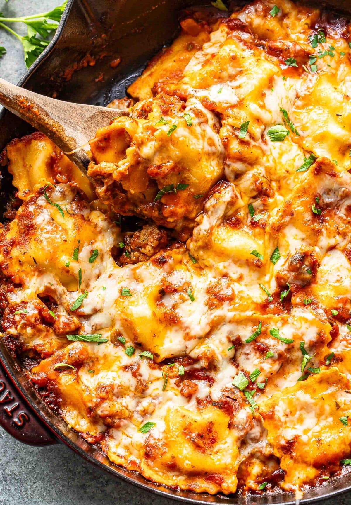 Wooden serving spoon scooping up baked ravioli out of a skillet.