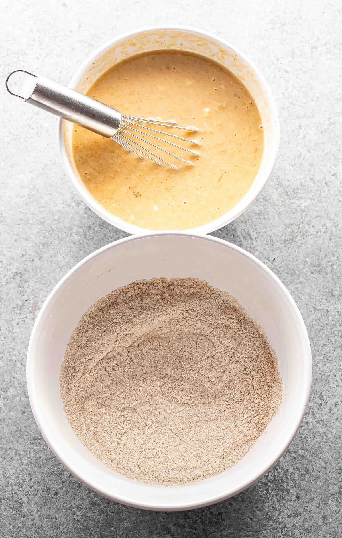 Dry ingredients for banana muffins in white mixing bowl and wet ingredients with a whisk in another mixing bowl.