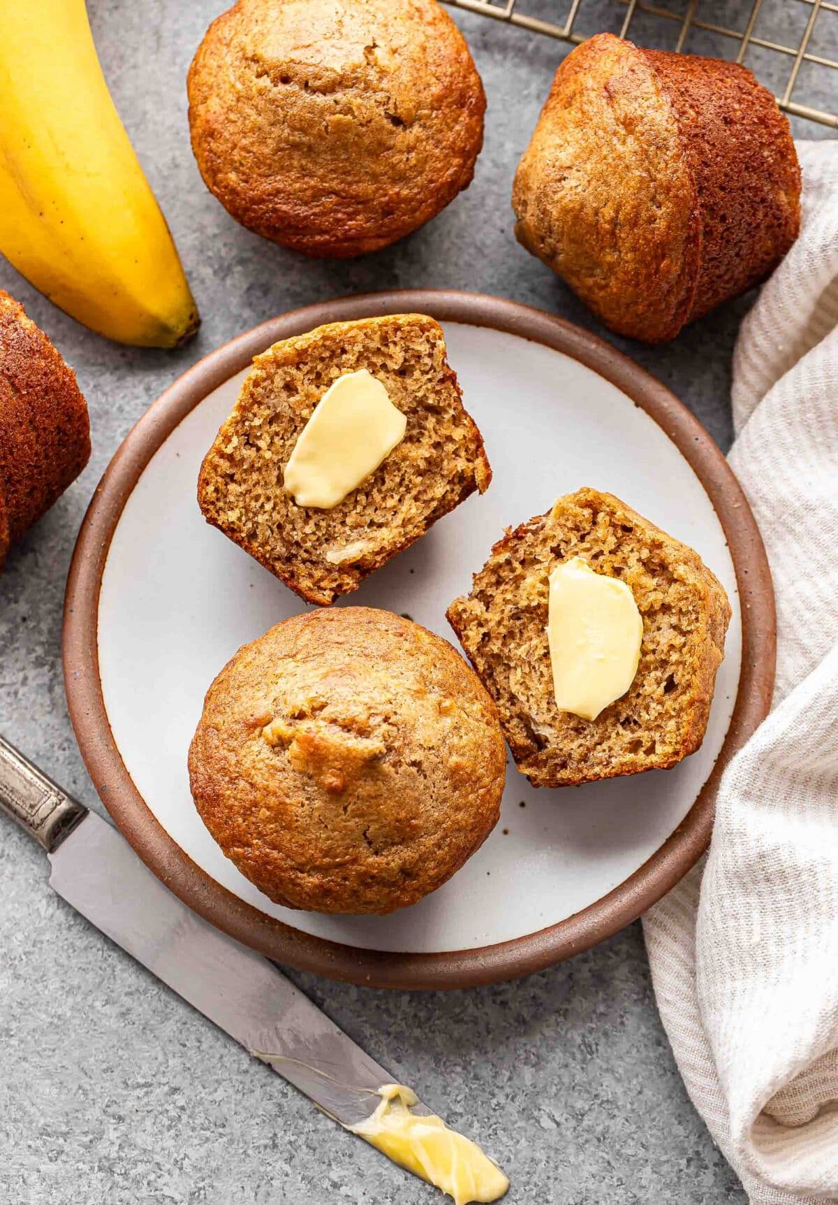 A whole banana muffin and one cut in half with butter on it on a white plate.