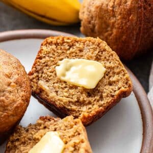 A banana muffin that's cut in half with butter on it on a white plate.