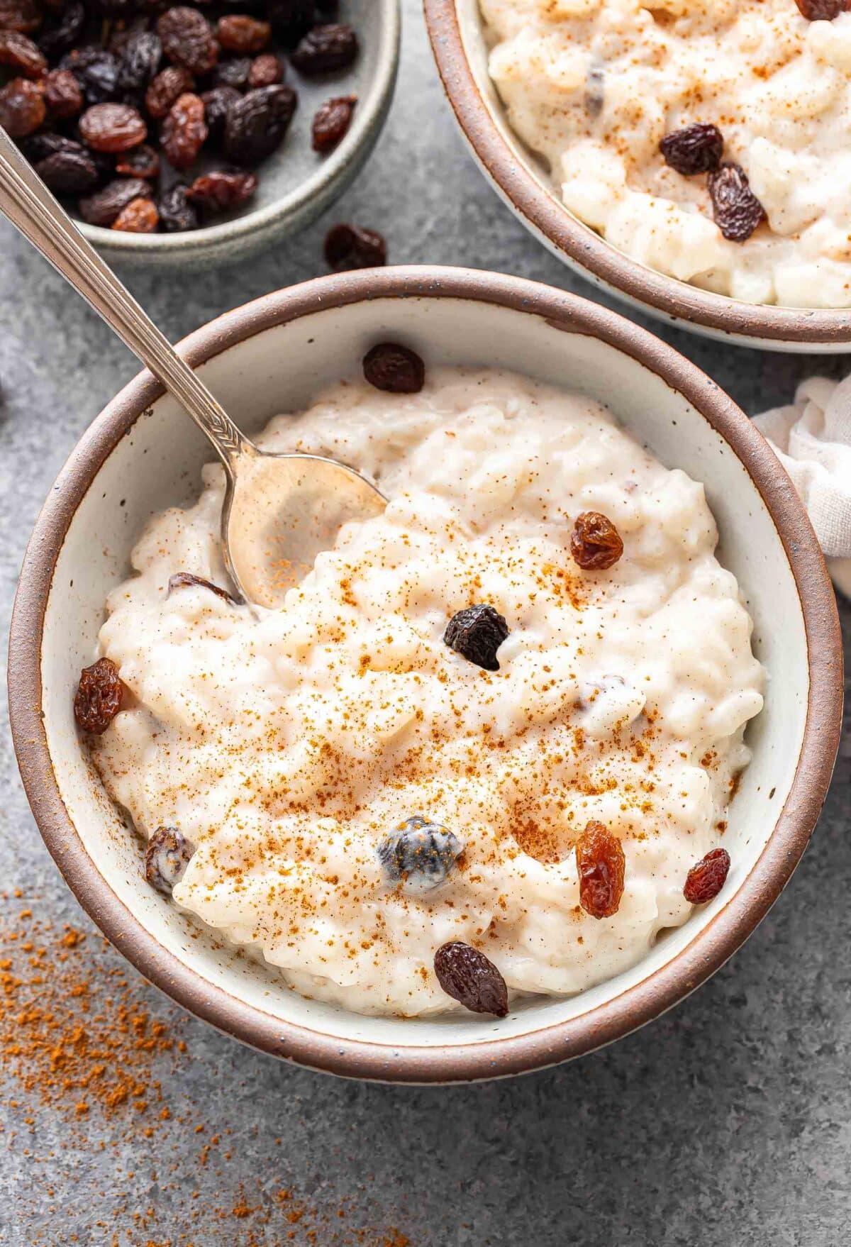 Rice pudding with raisins in a white bowl with a spoon. A bowl of raisins is behind the pudding.