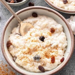 Rice pudding with raisins in a white bowl with a spoon. A bowl of raisins is behind the pudding.