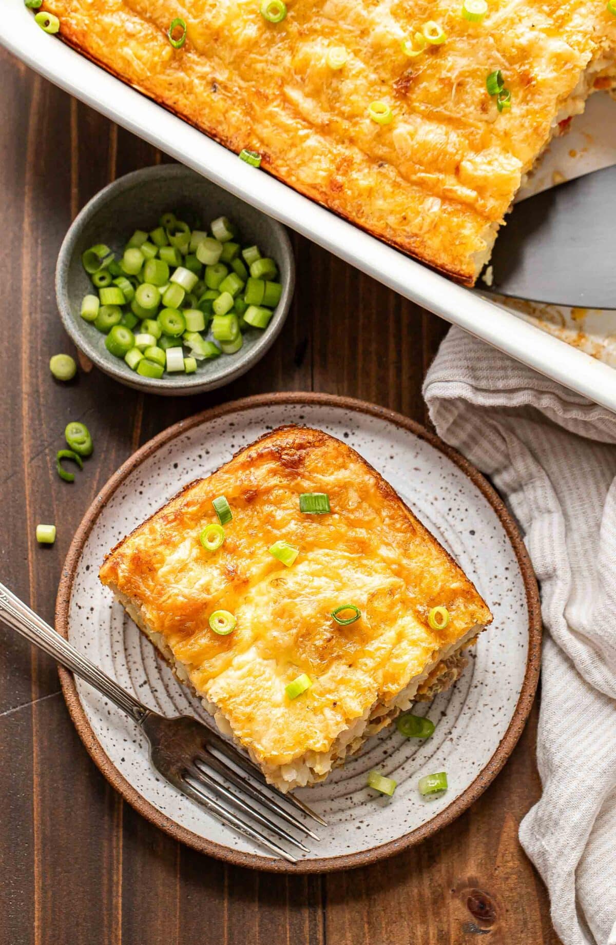 A piece of the egg breakfast casserole on a white plate with a fork and a bowl of sliced green onions behind it.