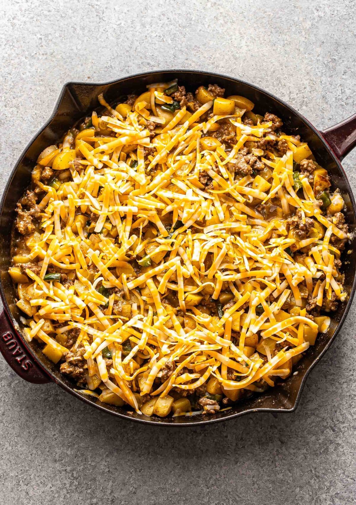 Ground beef potato casserole topped with shredded cheese in a skillet before being baked.