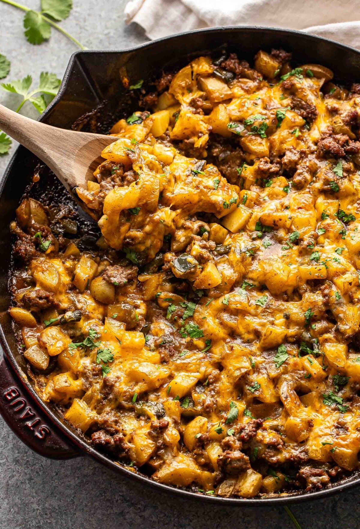 Ground beef potato casserole topped with melted cheese in a skillet with a wooden serving spoon.