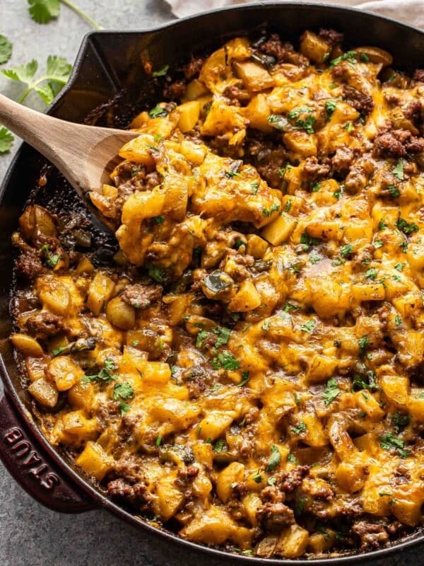 Ground beef potato casserole topped with melted cheese in a skillet with a wooden serving spoon.