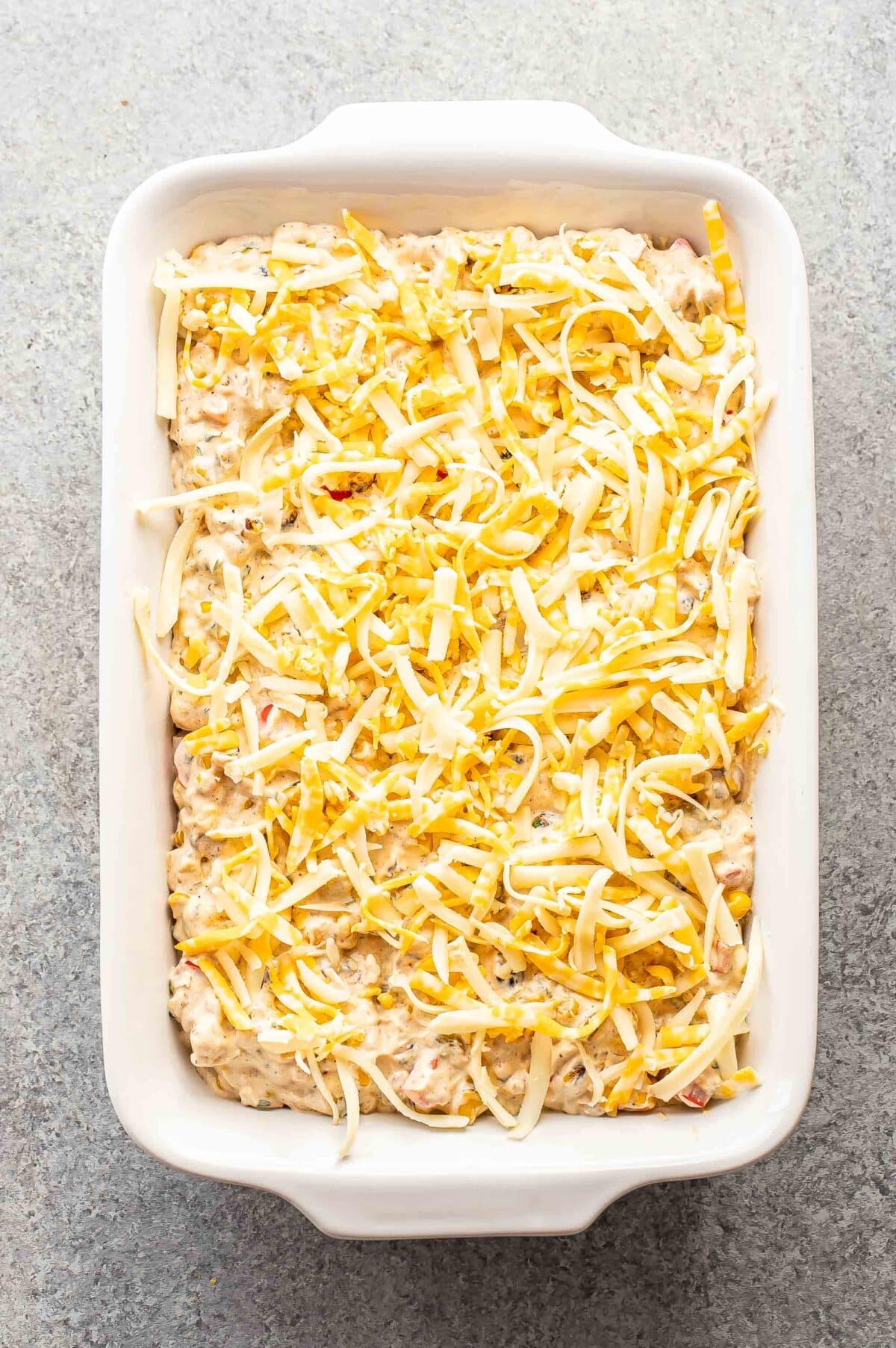 Creamy corn dip topped with shredded cheese in a white casserole dish before it's been baked.