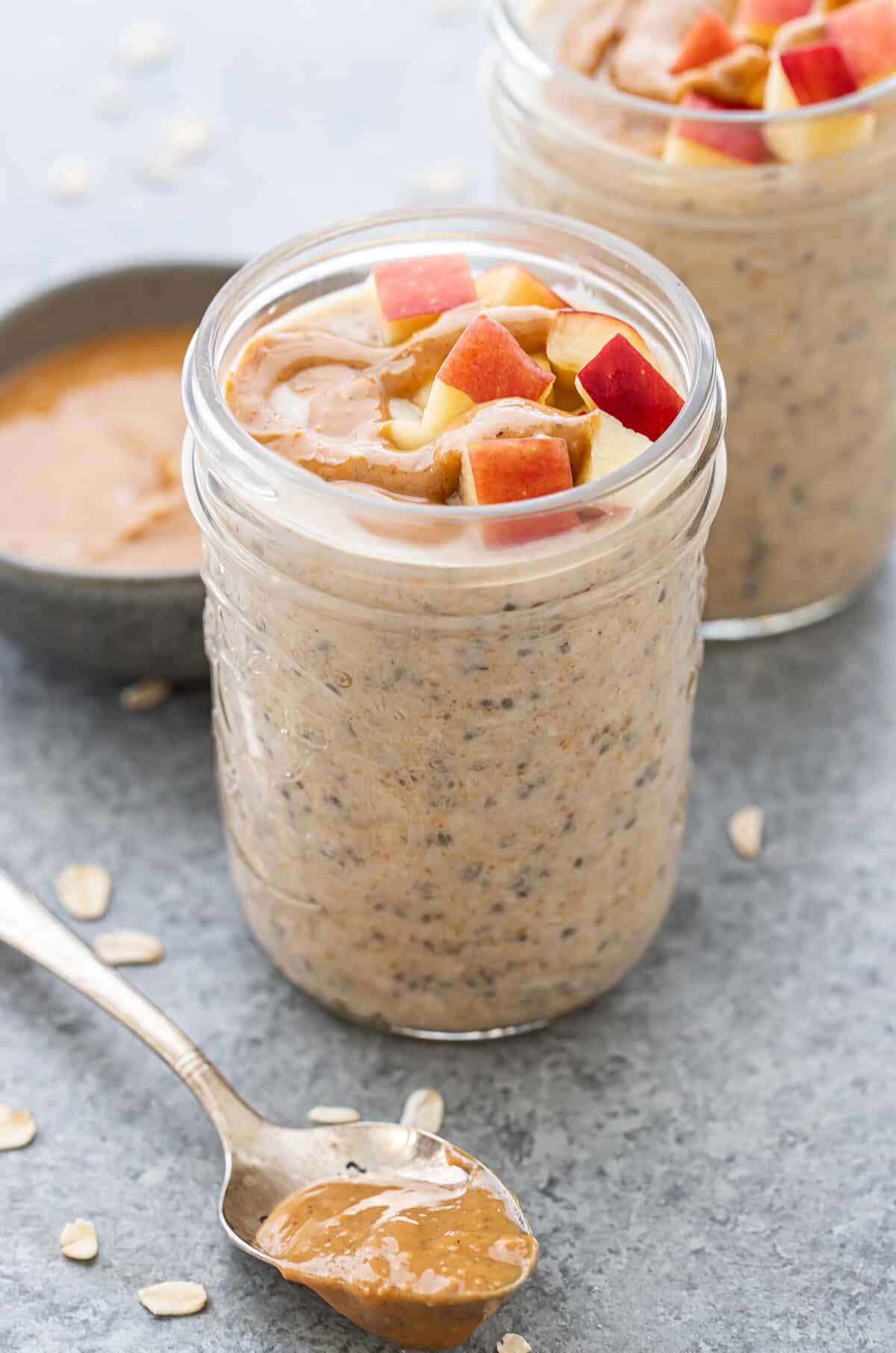 A jar of peanut butter overnight oats topped with chopped apples. A spoon of peanut butter is in front of the jar.