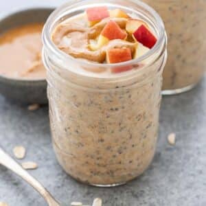 A jar of peanut butter overnight oats topped with chopped apples. A spoon of peanut butter is in front of the jar.