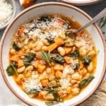 White bean soup with veggies in a white bowl with a spoon. Slices of crusty bread are next to the bowl.
