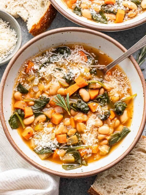 White bean soup with veggies in a white bowl with a spoon. Slices of crusty bread are next to the bowl.