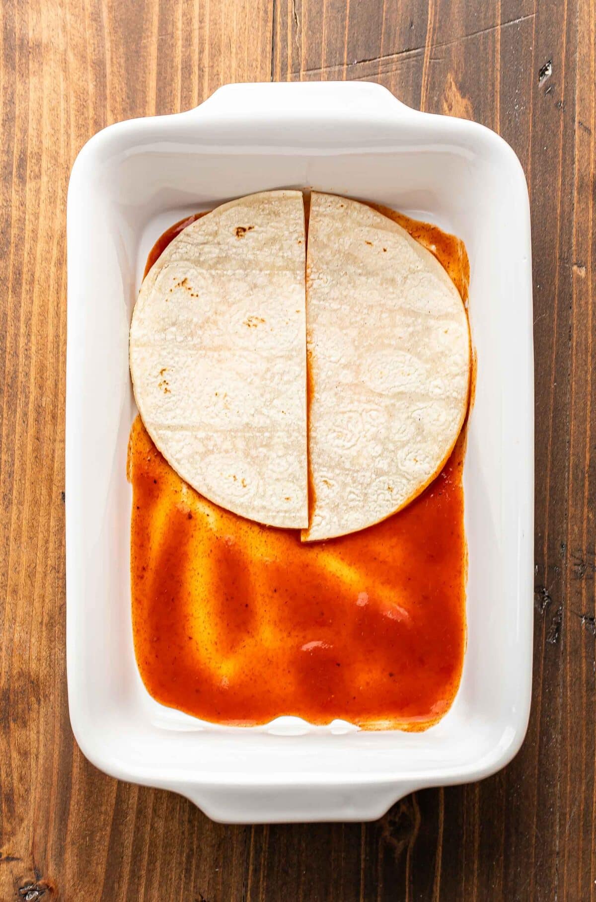 Casserole dish with red enchilada sauce and a corn tortilla in it.