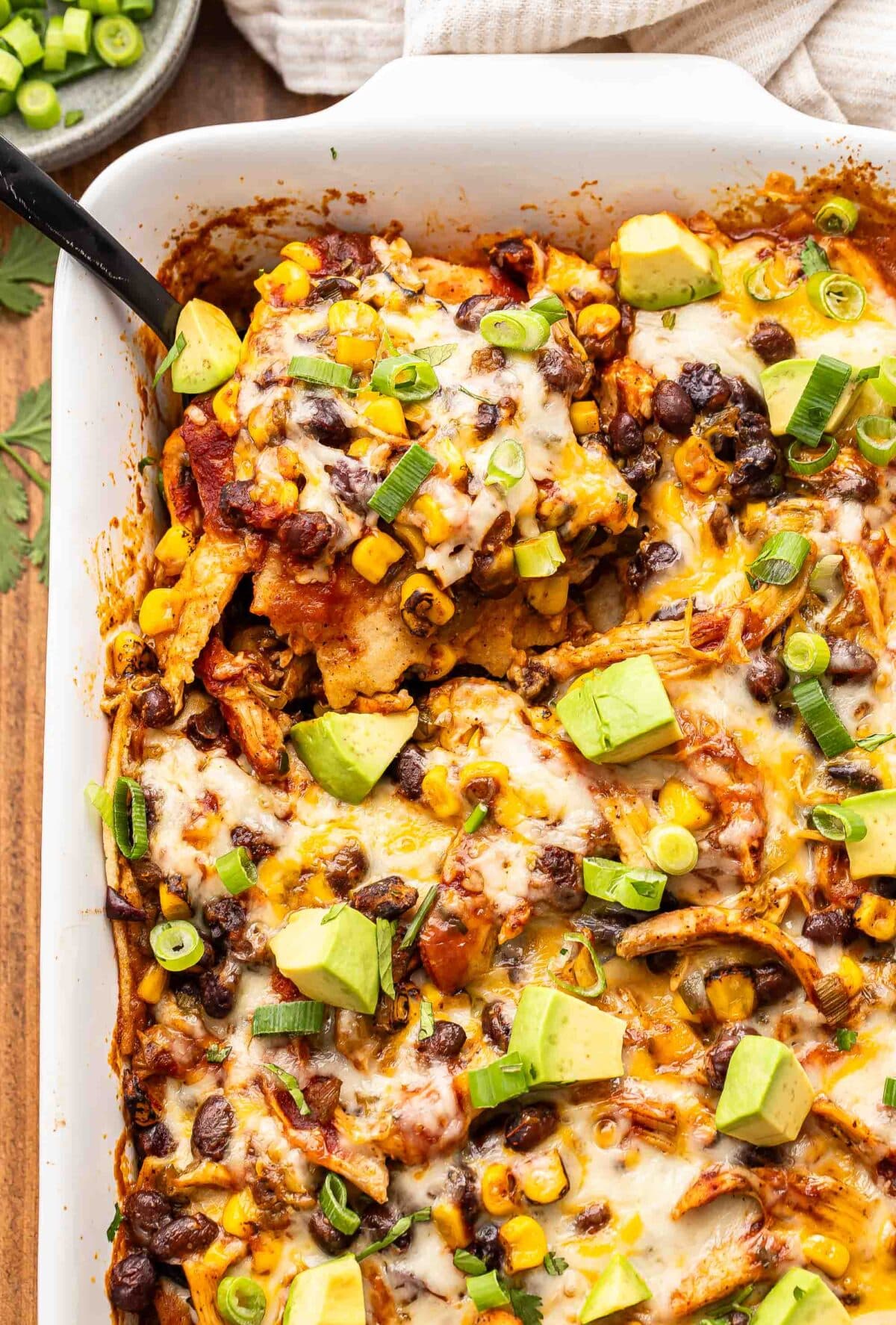 Black serving spoon scooping up chicken enchilada casserole out of the white baking dish.