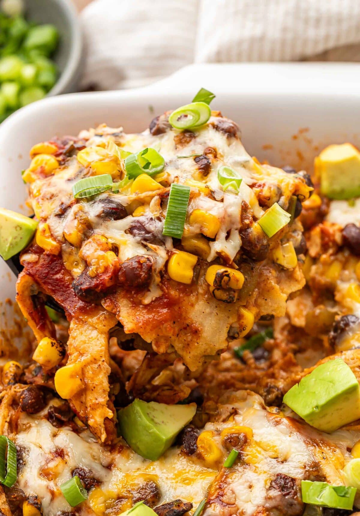 Serving spoon scooping up chicken enchilada casserole out of the white baking dish.