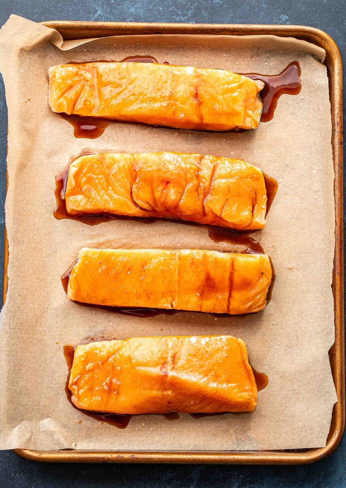 Four salmon fillets brushed with maple glaze on a parchment paper lined sheet pan before they've been baked.