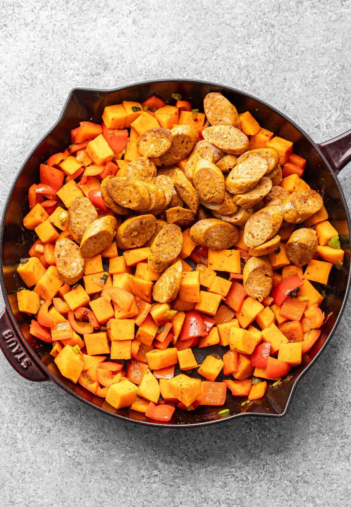 Cooked red bell peppers, sweet potato cubes, and green onion in a skillet with sliced sausage on top.