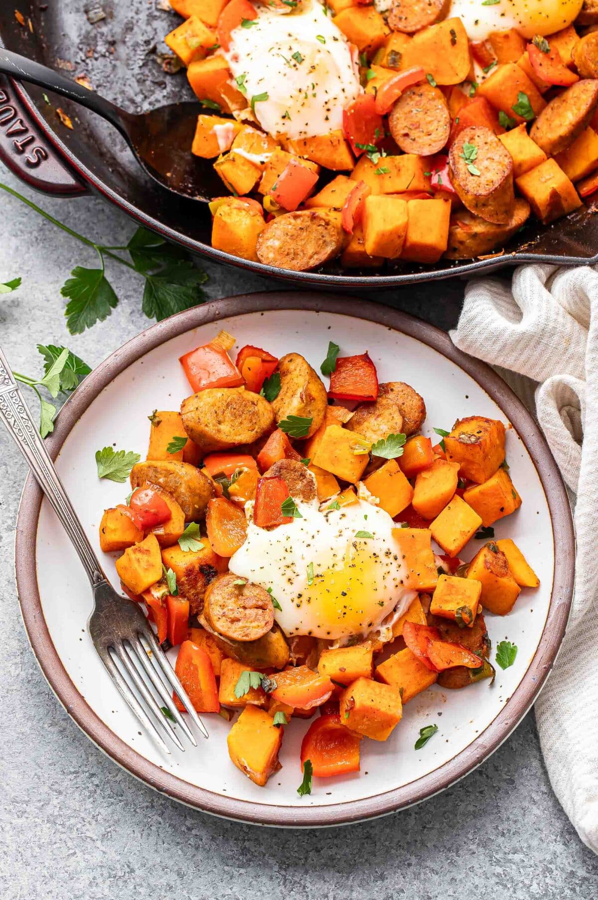 Sweet potato hash topped with an egg on a white plate with a fork. The skillet of hash is behind the plate.