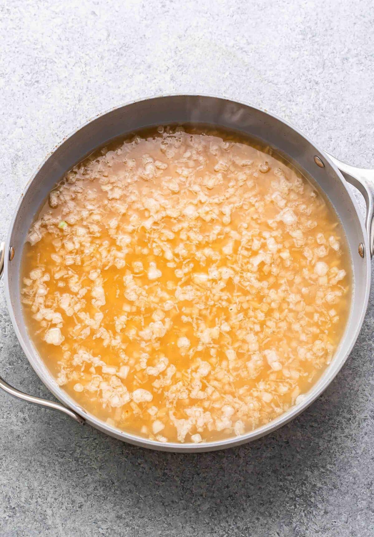 Orzo, onion, garlic, lemon zest, and chicken broth in a skillet before it's been cooked.