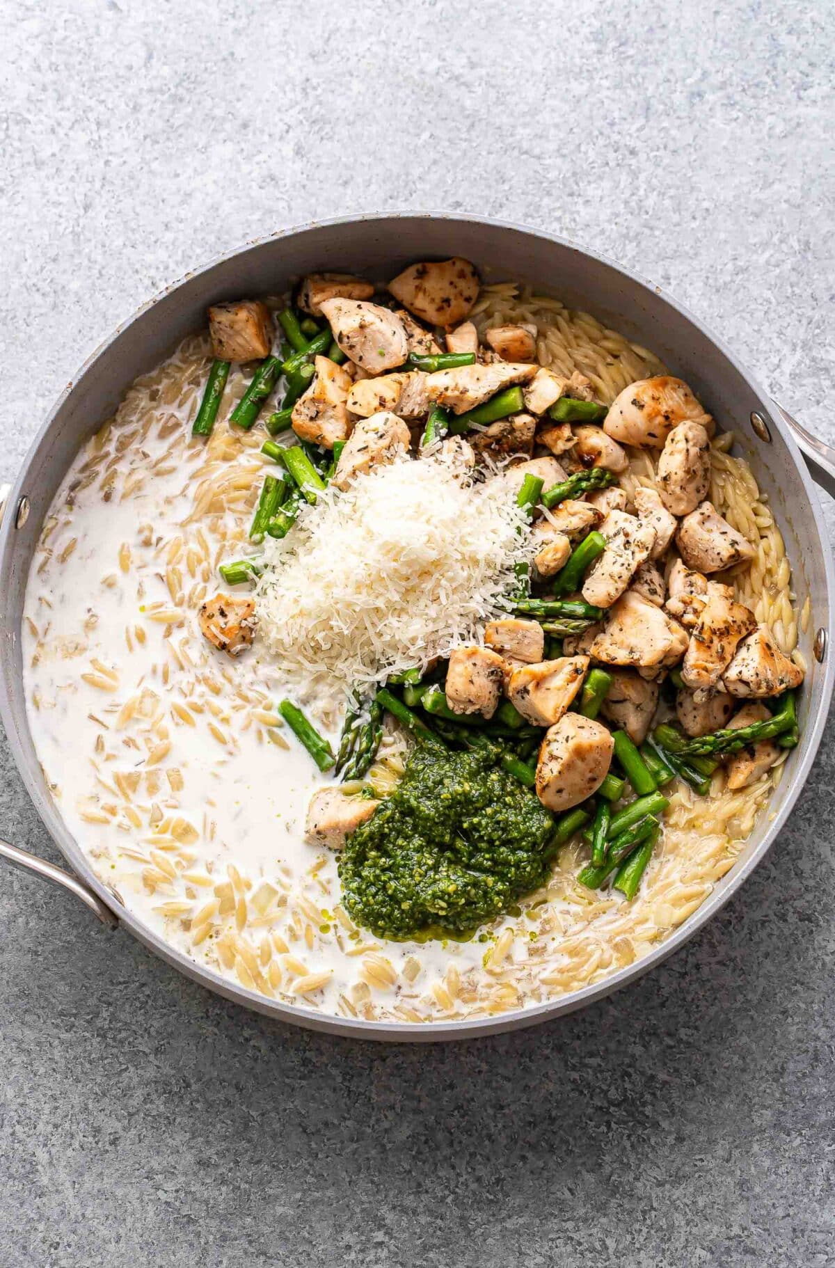 Cooked orzo in a pan with cooked chicken, asparagus, pesto, cream, and parmesan cheese on top before they've been stirred in.