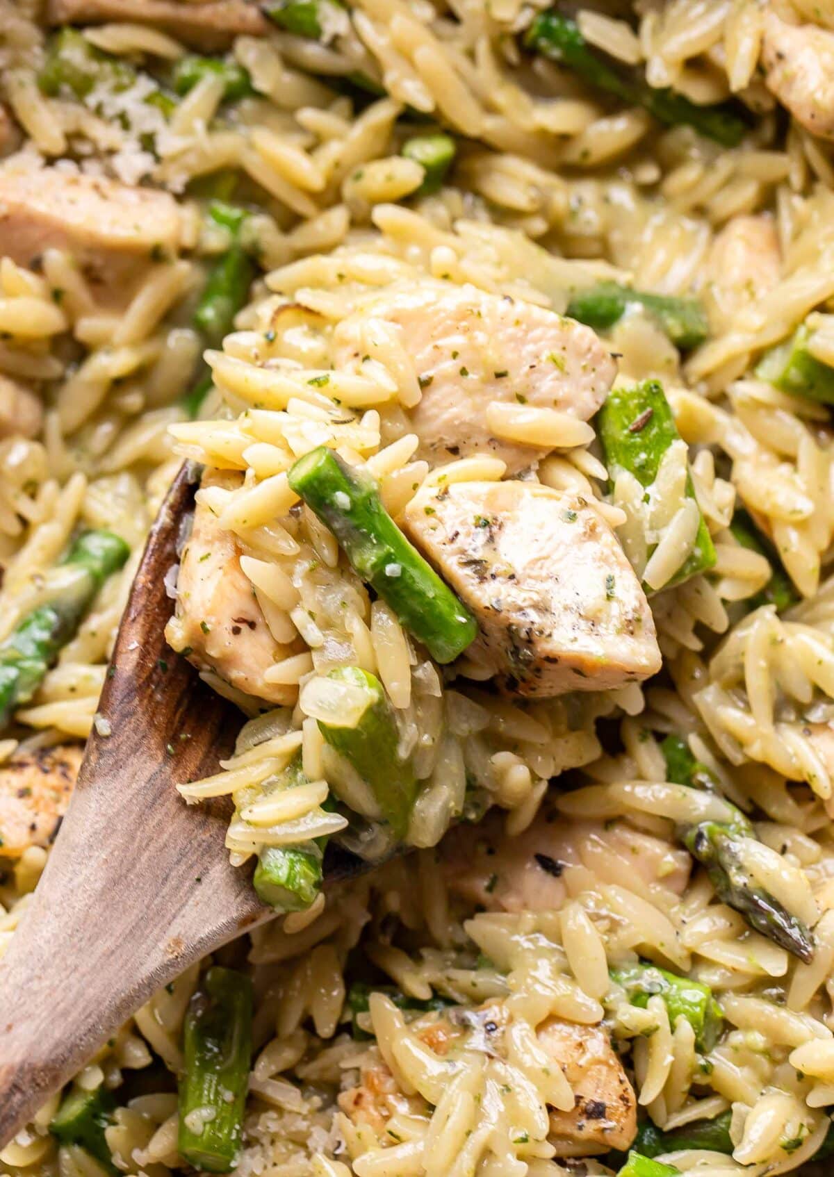 Wooden serving spoon scooping up asparagus orzo with chicken out of a pan.