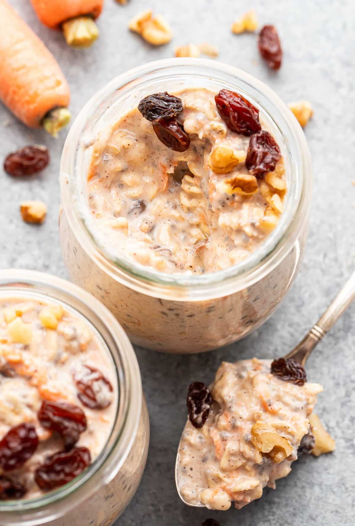 Two jars of carrot cake overnight oats topped with raisins and walnuts. A spoonful of the oats is laying next to one of the jars.