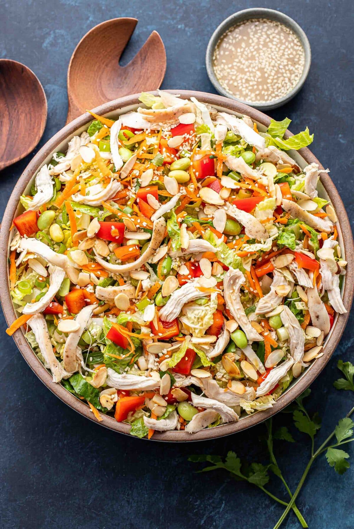 Chinese chicken salad in a white salad bowl with wooden serving spoons and a bowl of dressing behind it.