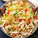 Asian chicken salad in a white salad bowl with wooden serving spoons in the bowl and dressing behind it.